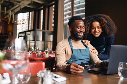 Business Couple holding an APGFCU Visa Business Cash Back Card making purchases on computer